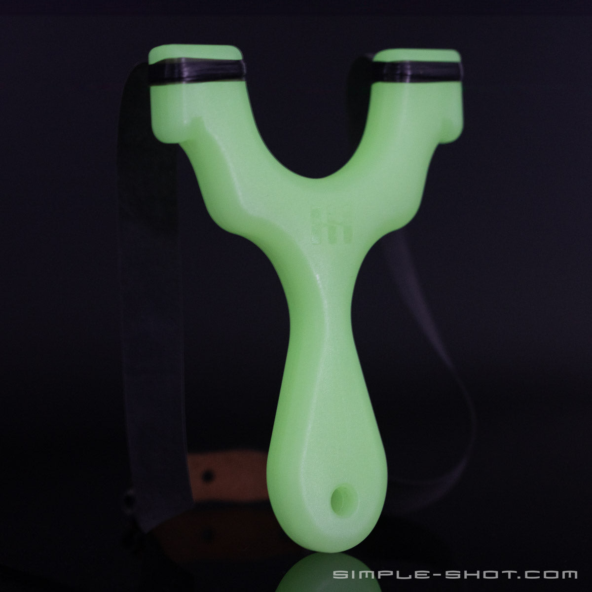 A Glow-in-the-Dark Axiom Champ Slingshot with a sleek, modern design, featuring a Y-shaped frame and black elastic bands. The handle boasts a textured grip for superior hold, and the minimalist gear brand name "Simple-Shot.com" is visible at the bottom right corner of the image.