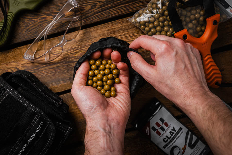 Hands cupping a pile of clay slingshot ammo over a mesh bag, with a slingshot and clay packaging on a wood table